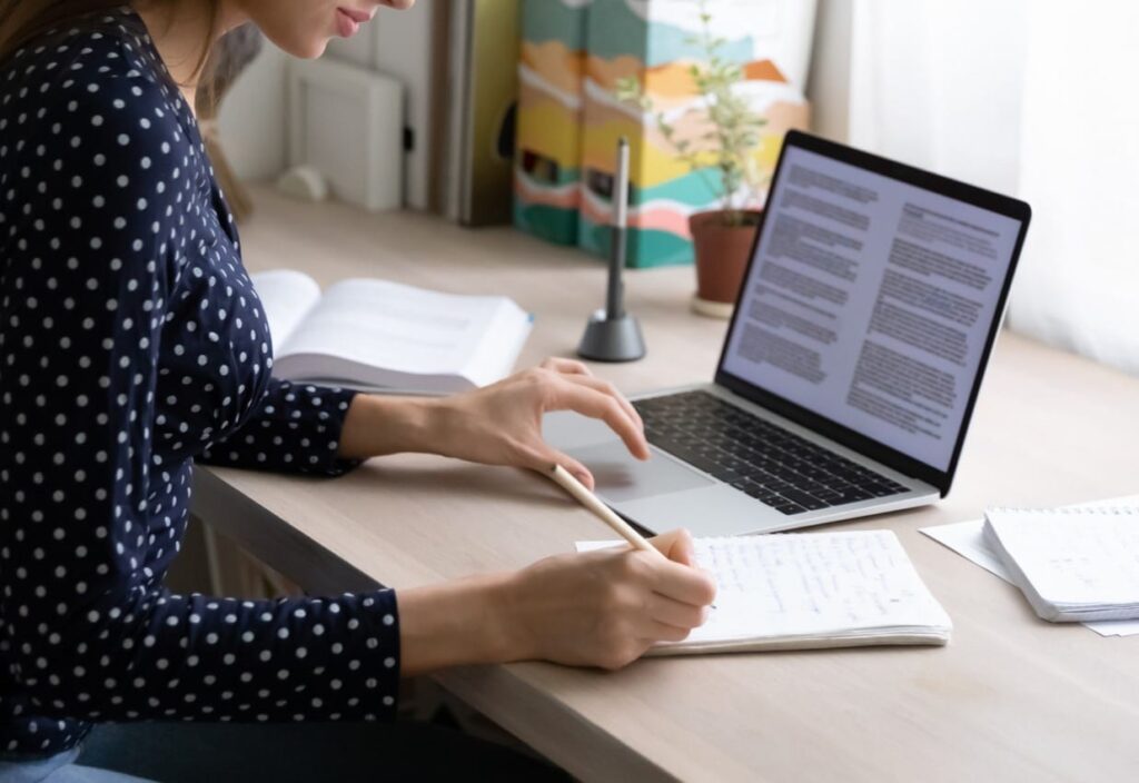 Eine Frau in blauer Bluse macht sich Notizen auf Papier und arbeitet zugleich an einem Laptop