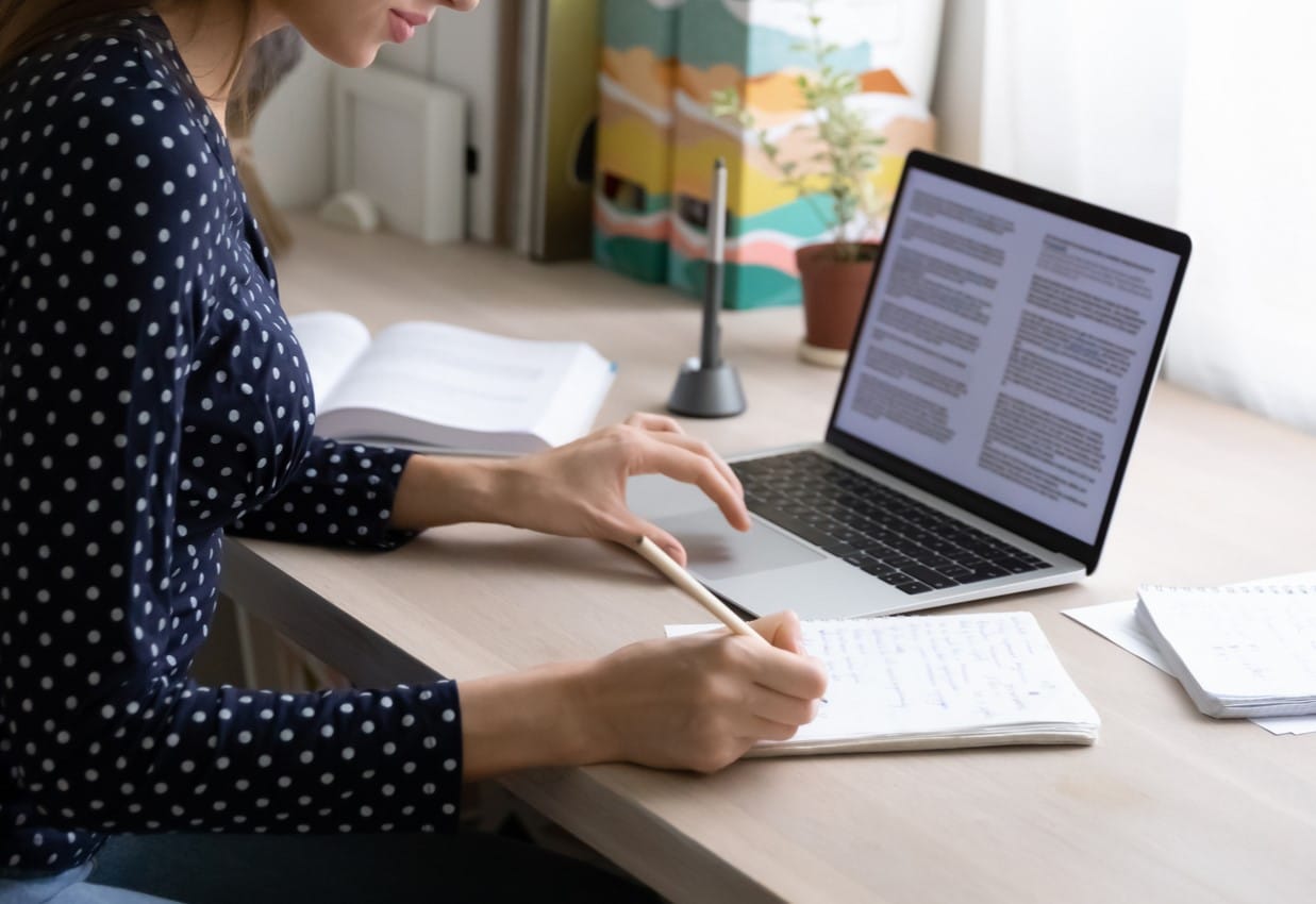 Eine Frau in blauer Bluse macht sich Notizen auf Papier und arbeitet zugleich an einem Laptop