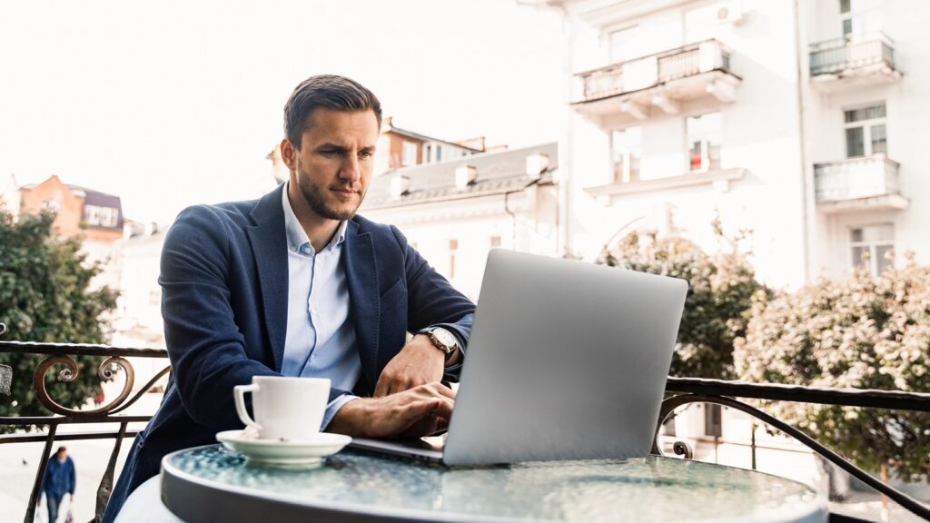 Geschäftsmann in einer blauen Jacke arbeitet an einem Laptop auf der Terasse eines Cafés. Neben ihm steht eine Tasse Kaffee.