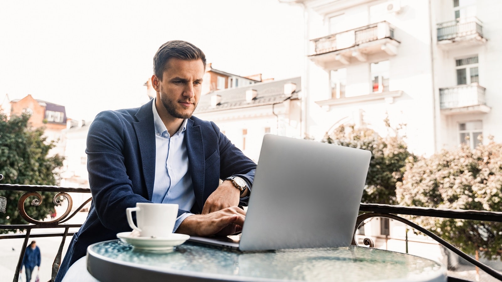Geschäftsmann in einer blauen Jacke arbeitet an einem Laptop auf der Terasse eines Cafés. Neben ihm steht eine Tasse Kaffee.