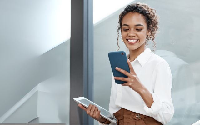 Frau mit Tablet in der einen Hand schaut lächelnd auf Smartphone