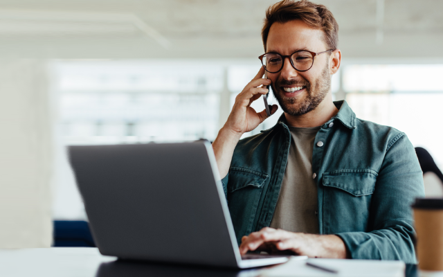 Mann mit Brille sitzt vor Laptop und telefoniert am Handy