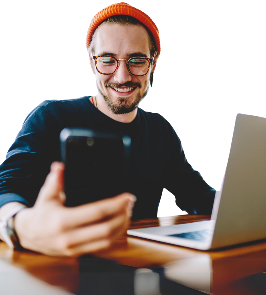 Ein Mann sitzt vor einem Laptop und sieht parallel auf sein Smartphone, das er in seiner Hand hält.