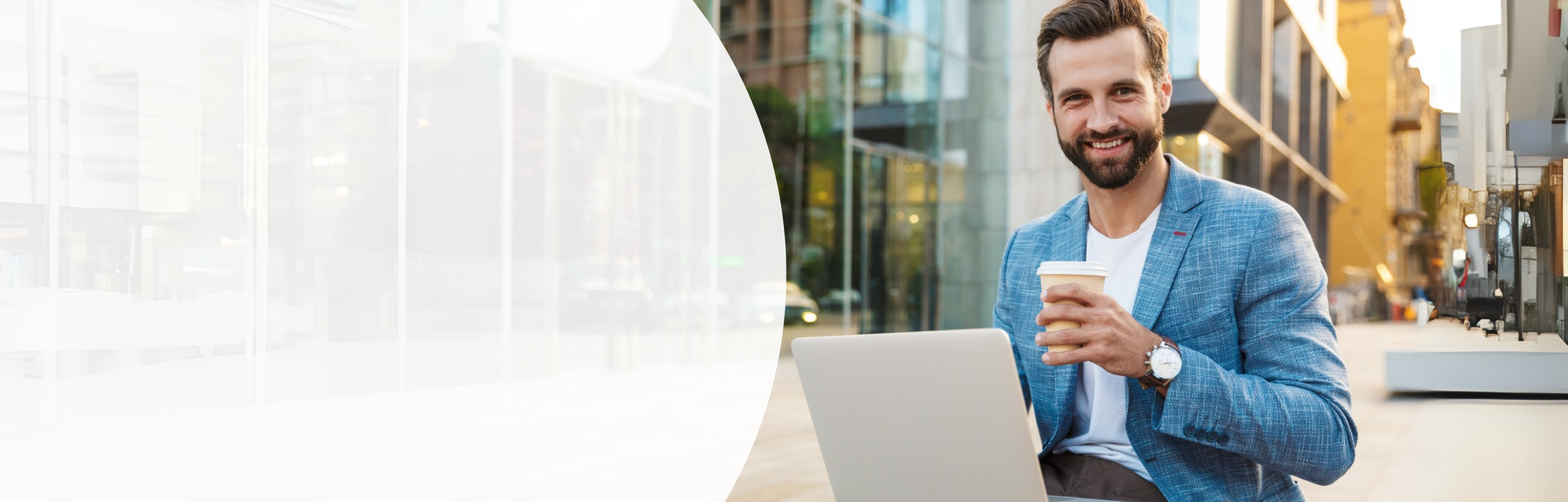 Mann mit Bart sitzt an einem öffentlichen Platz vor seinem Laptop und hält einen Kaffeebecher in der Hand