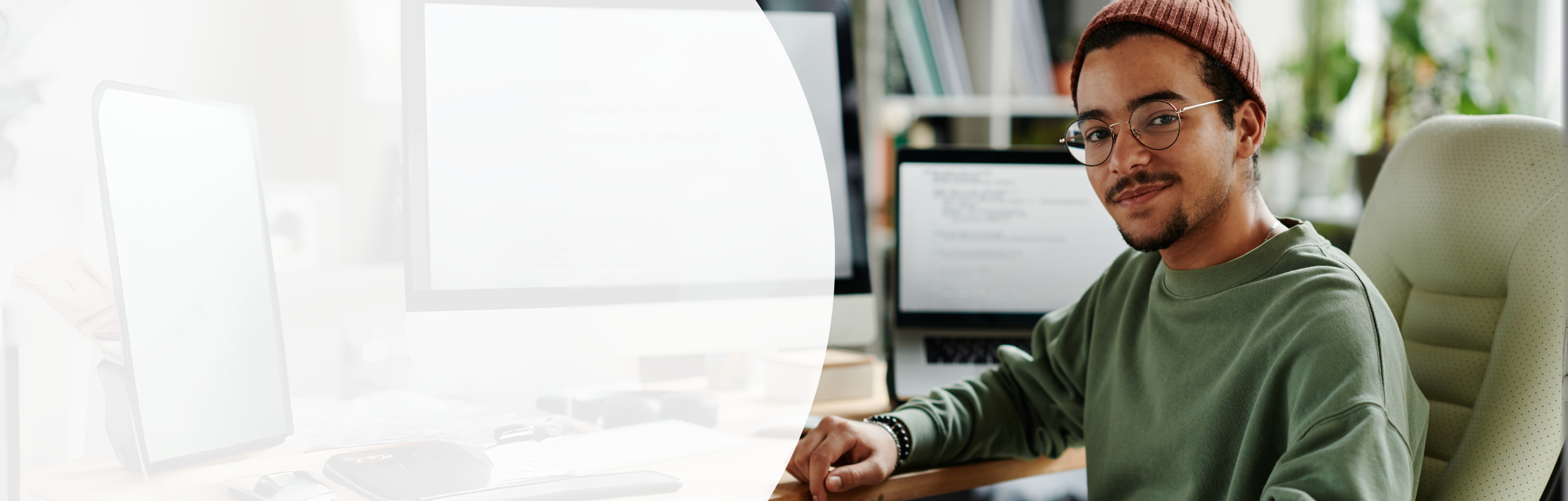 Ein Mann in einem dunkelgrünen Pullover und Mütze sitzt an einem Bildschirmarbeitsplatz