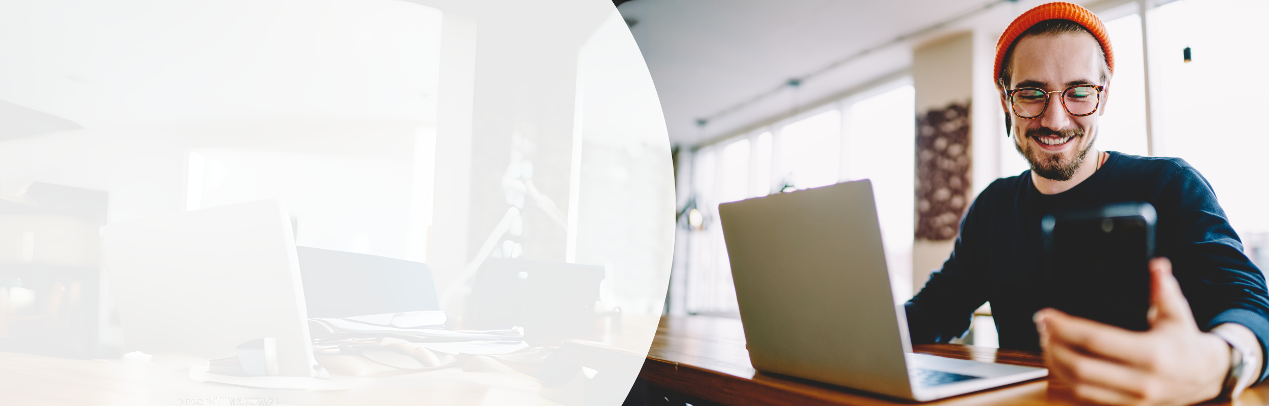 Mann mit orangener Mütze und Brille sitzt vor Laptop und schaut lächelnd auf sein Handy