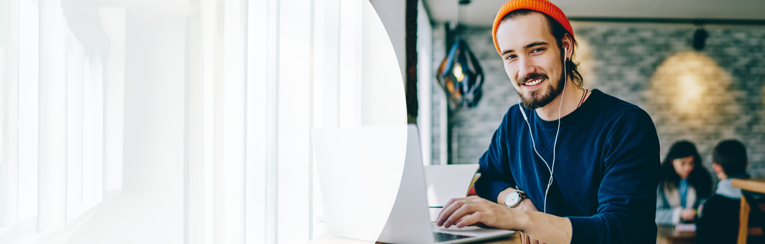 Mann mit Bart, Earphones und orangener Mütze sitzt am Laptop und lächelt