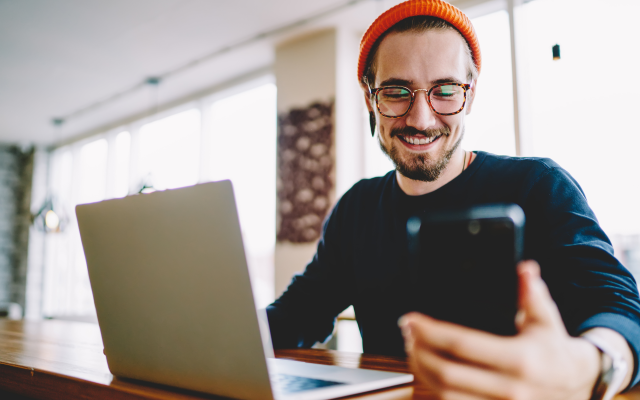 Mann mit orangener Mütze und Brille sitzt vor Laptop und schaut lächelnd auf sein Handy