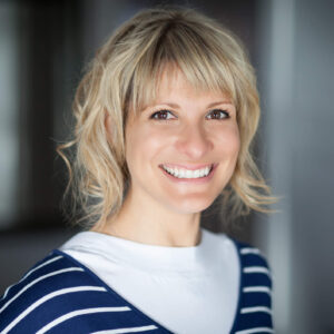 Portrait einer blonden Dame, die ein weißes Shirt und eine blaugestreifte Weste trägt.