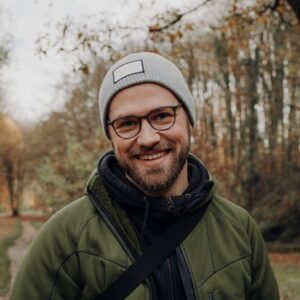 Portrait eines jungen lächelnden Mannes, der eine Brille trägt und eine hellgraue Mütze.