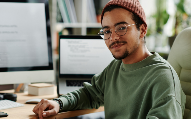 Ein Mann in einem dunkelgrünen Pullover und Mütze sitzt an einem Bildschirmarbeitsplatz