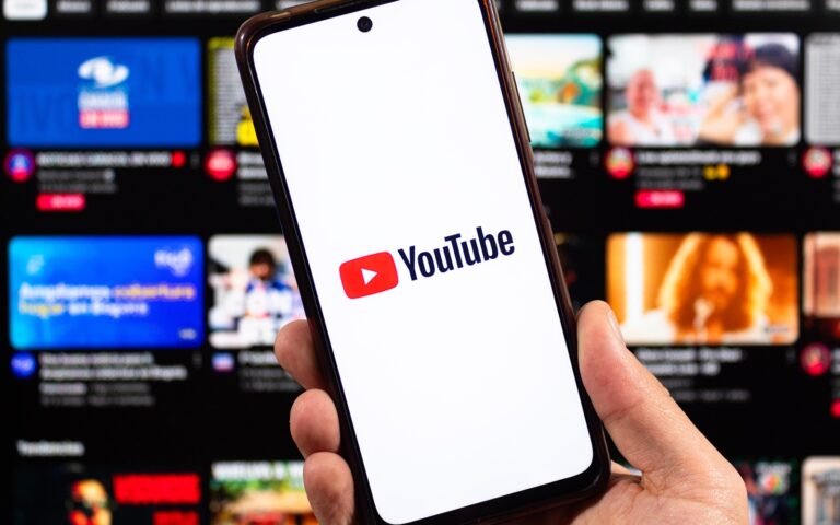 Eine Hand, die ein Smartphone hält, auf der ein Youtube-Logo abgebildet ist. Mit verschwommener Youtube-Benutzeroberfläche im Hintergrund.